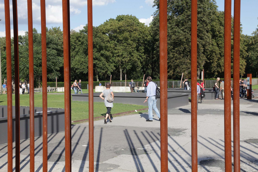 Das Außenareal der Gedenkstätte Berliner Mauer im Sommer. Zu sehen sind vertikale Eisenstäbe, welche den Mauerverlauf kennzeichnen und grüner Rasen.