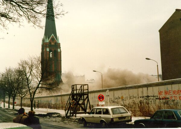 Die Berliner Mauer ist zu sehen mit einigen Graffitis und Autos an der Bernauer Straße. Hinter der Mauer sieht man den Kirchenturm und Staub in der Luft, der von der Sprengung kommt. 