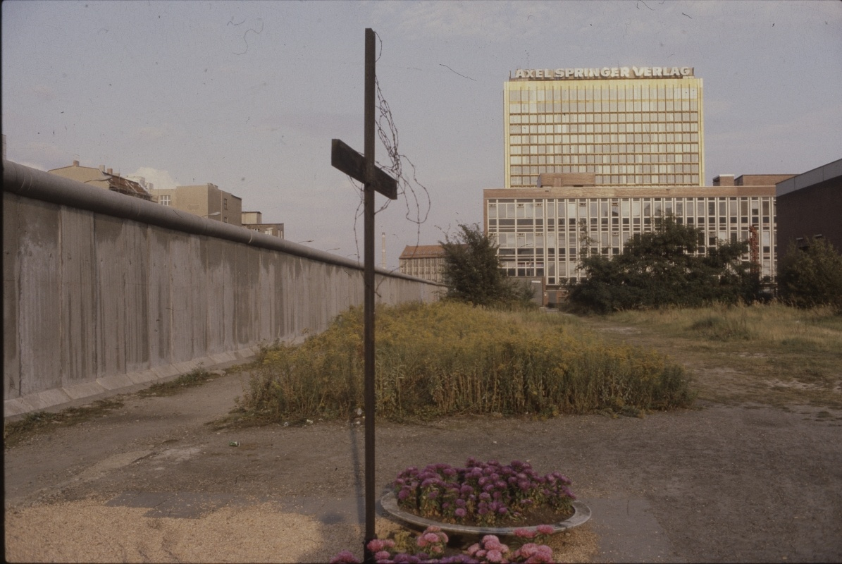 Kreuz steht neben der Berliner Mauer, im Hintergrund ist ein Hochhaus zu sehen