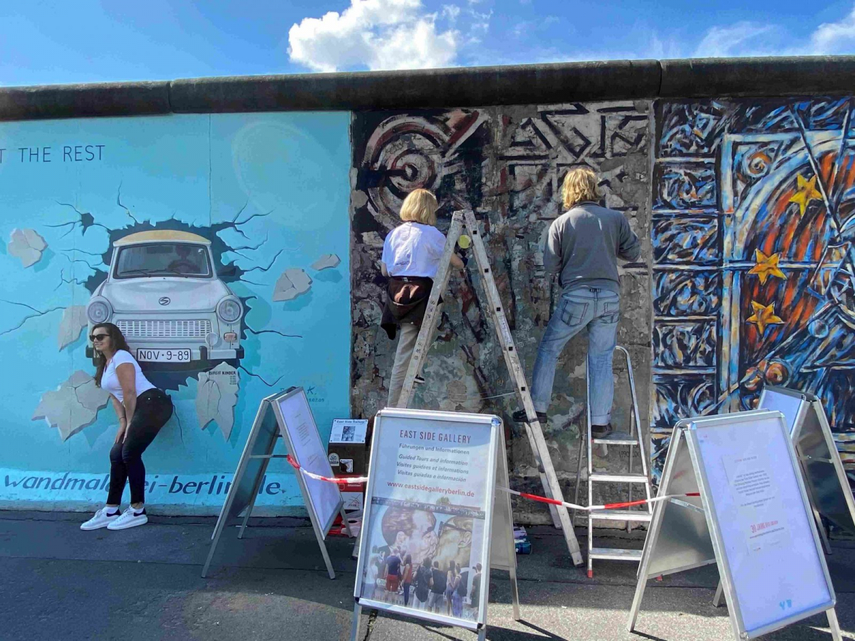 Zwei Personen konservieren ein Bild an der East Side Gallery und daneben posiert eine Frau für ein Foto
